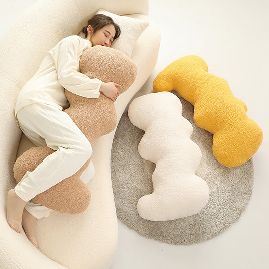 Three cloud-shaped fuzzy cushions in beige, cream and mustard, one hugged by a woman on a sofa.
