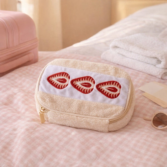 Strawberry sando makeup bag on a pink gingham bed beside a pink suitcase, packed for a trip