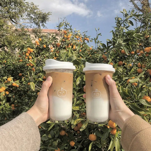 Two clear cups with white lids and apple face print, filled with iced latte in an orange grove
