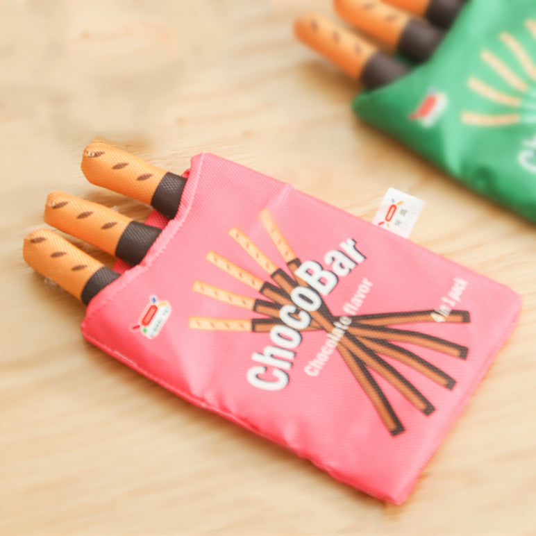 One pink pouch labelled ChocoBar holding three orange-brown chocolate-tipped stick plushes on a wood surface.