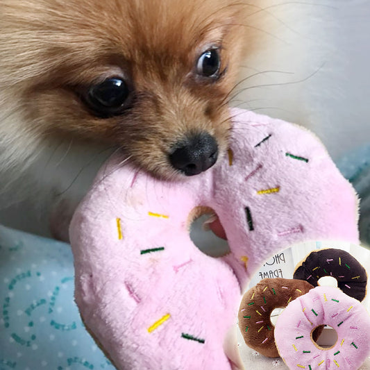 Small tan Pomeranian biting a pink plush donut with embroidered multicolor sprinkles.