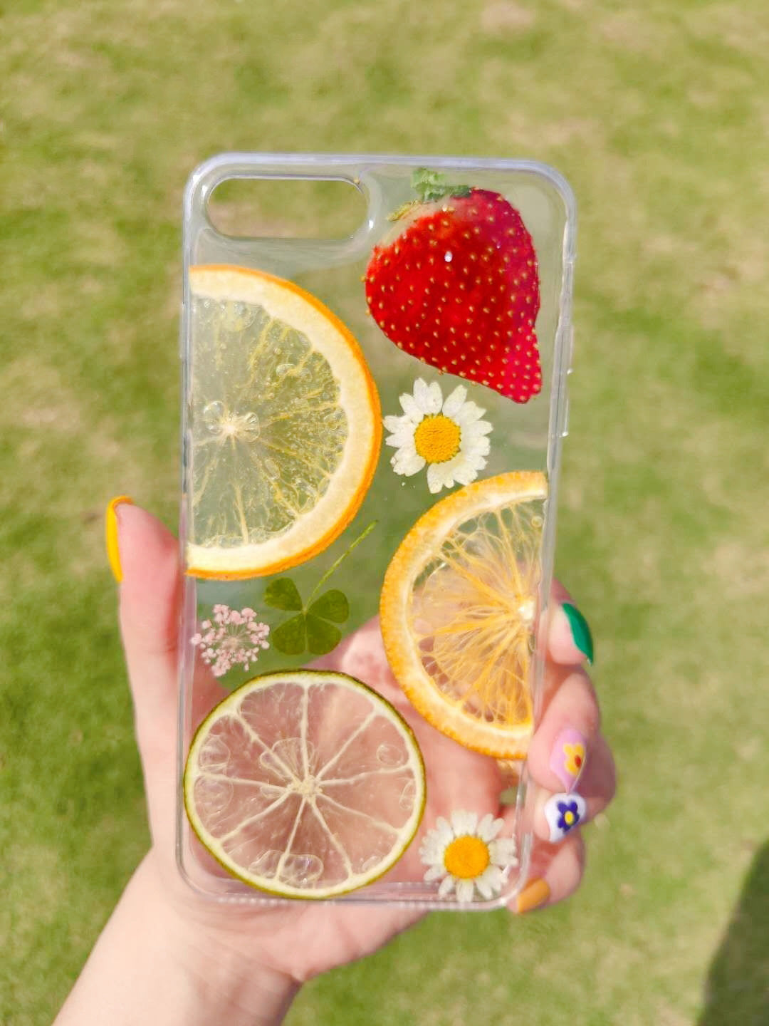 Clear iPhone case with embedded dried fruit slices — orange, lime, and a strawberry, along with white flowers.