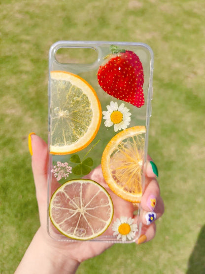 Clear iPhone case with embedded dried fruit slices — orange, lime, and a strawberry, along with white flowers.