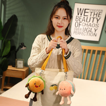Woman holding cream tote with yellow straps and two plush keychains: green burger and pink dessert.