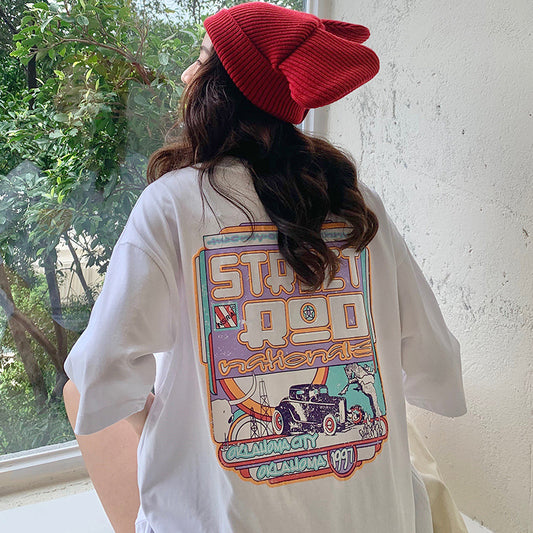 Back view of white oversized T-shirt with large colorful retro car and text graphic; red knit beanie visible.