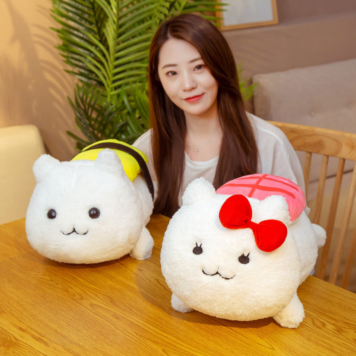 Two white cat plush toys on a wooden table, one with pink sushi top and red bow, one with yellow sushi top.