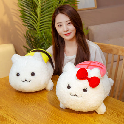 Two white cat plush toys on a wooden table, one with pink sushi top and red bow, one with yellow sushi top.