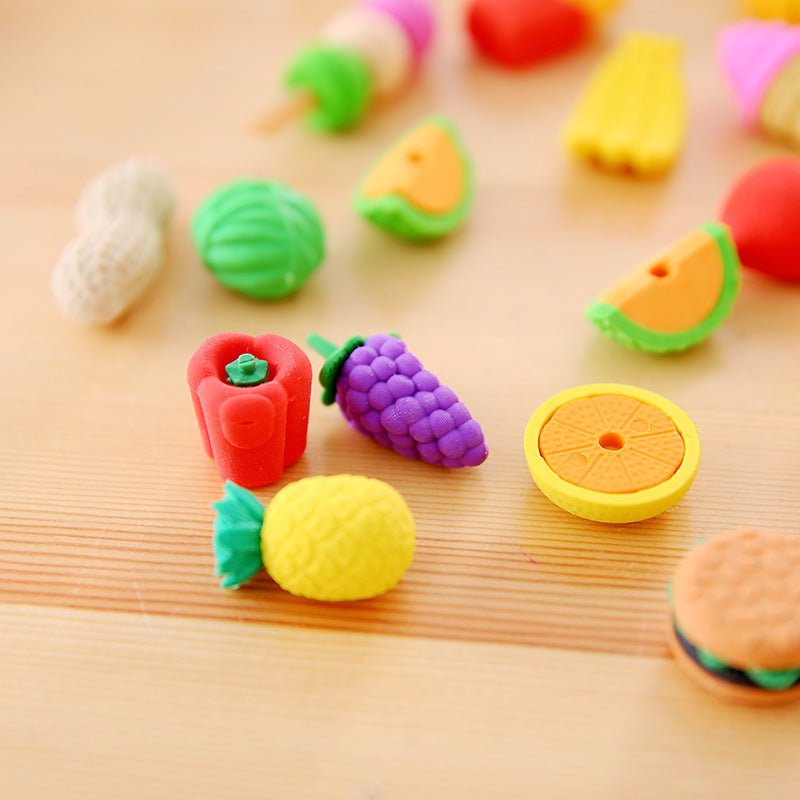 13 colorful food erasers on wood: pineapple, purple grapes, red pepper and an orange slice.