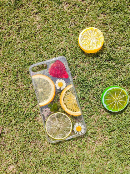 One clear iPhone case on grass with dried citrus slices, a strawberry, two daisies and two citrus halves.