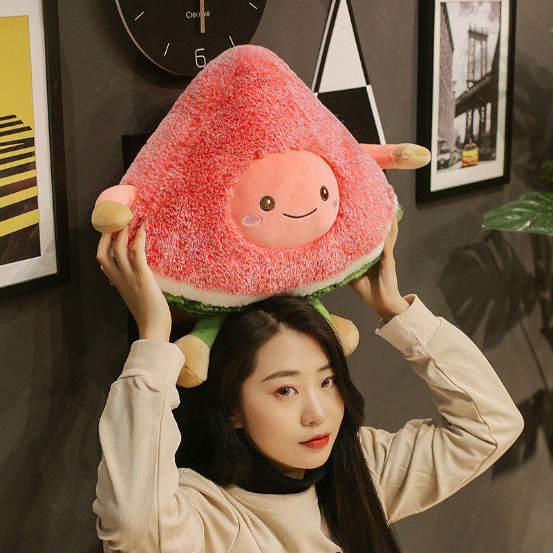 Woman holding a large triangular pink fuzzy fruit plush with smiling round face, green rind and stubby limbs.