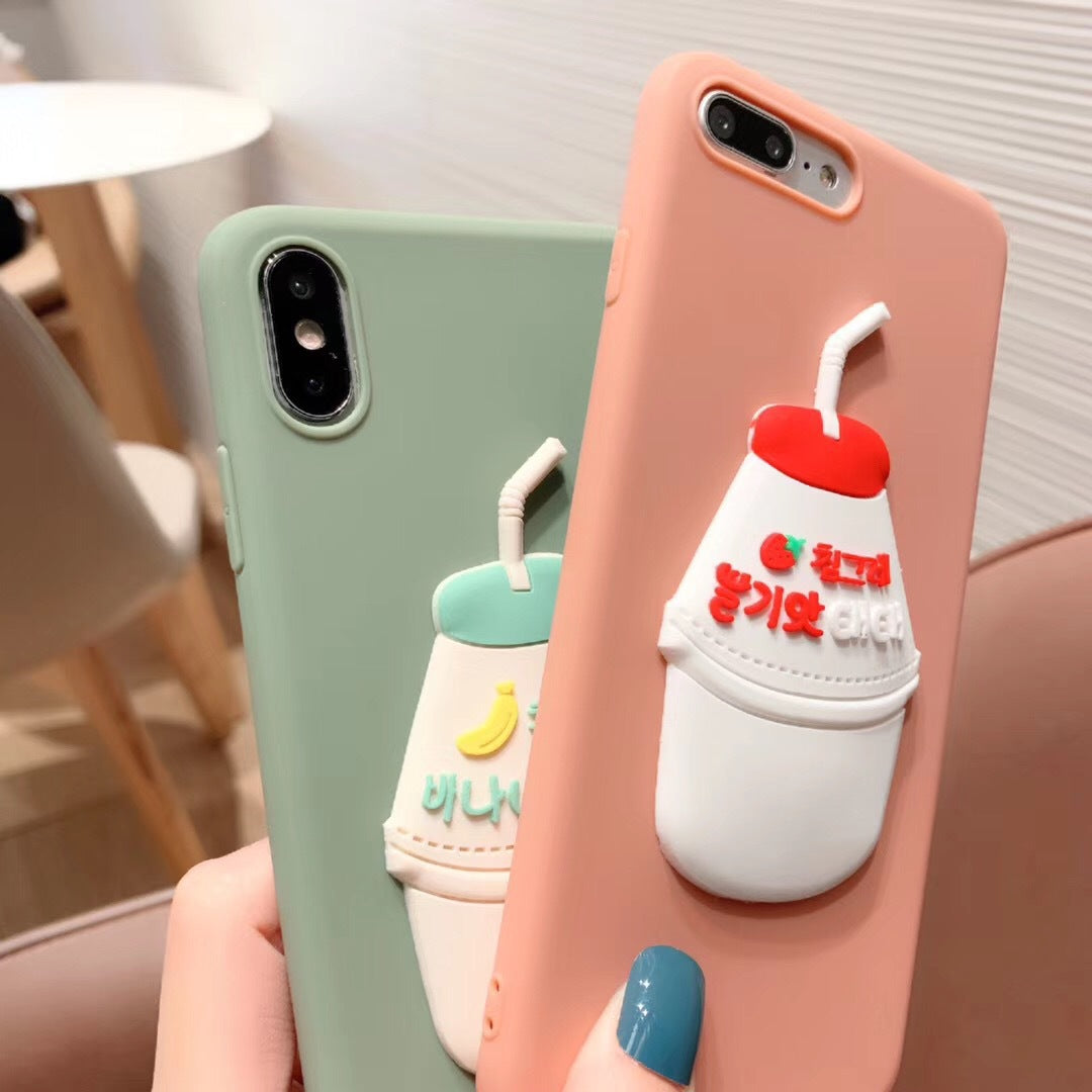 Hand holding two silicone cases: sage green with 3D banana milk charm, peach with 3D strawberry milk charm.
