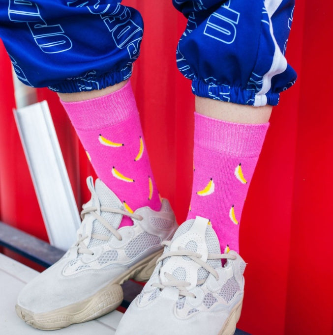 Bright pink ankle socks featuring playful banana designs, paired with light grey trainers and blue joggers.