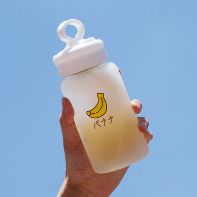 Hand holding one frosted bottle with white loop cap, pale yellow liquid and banana graphic and Japanese text.