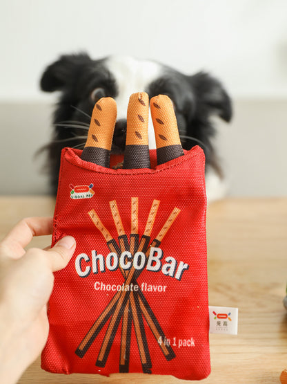 Hand holding one red ChocoBar fabric pouch with three orange-brown stick plushes, dog blurred behind.