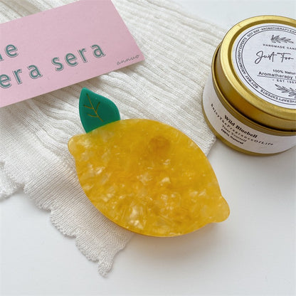 One yellow marbled lemon-shaped hair clip with a small green leaf, on white cloth beside a gold tin.