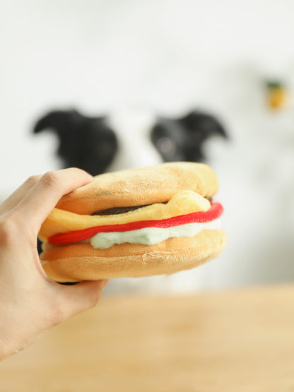 Hand holding one plush hamburger toy with tan buns, red stripe, green and yellow layers, and brown patty.