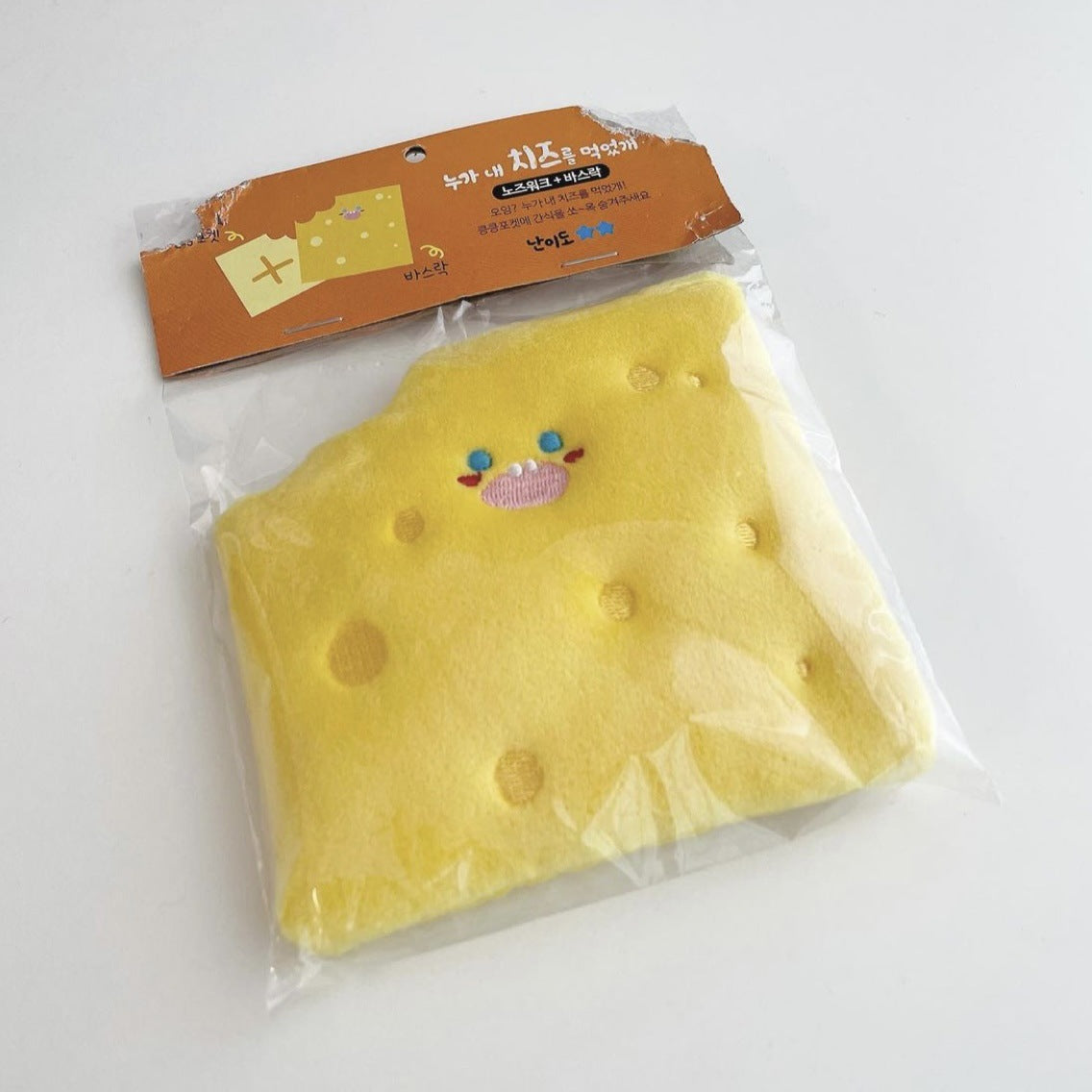Yellow cheese wedge dog toy with crinkle material and a cute face, packaged in transparent plastic.