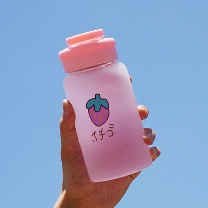 Hand holding one frosted pink bottle with wide lid, strawberry graphic and Japanese text against blue sky.