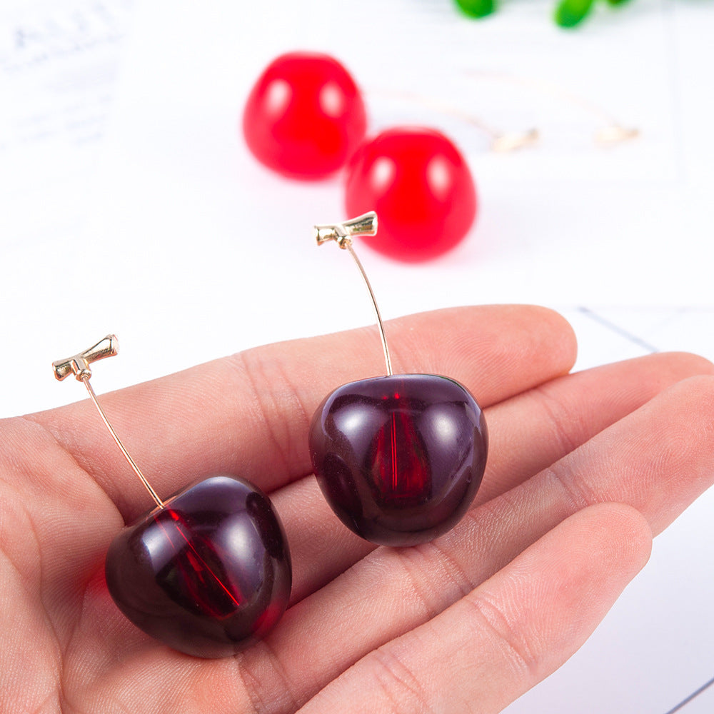 Cherry-shaped stud earrings in deep red with gold-tone connectors, resting on a hand, with another pair blurred in the bac...