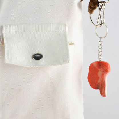 Silver key ring and chain holding one small realistic red steak charm hanging beside a white pocket.