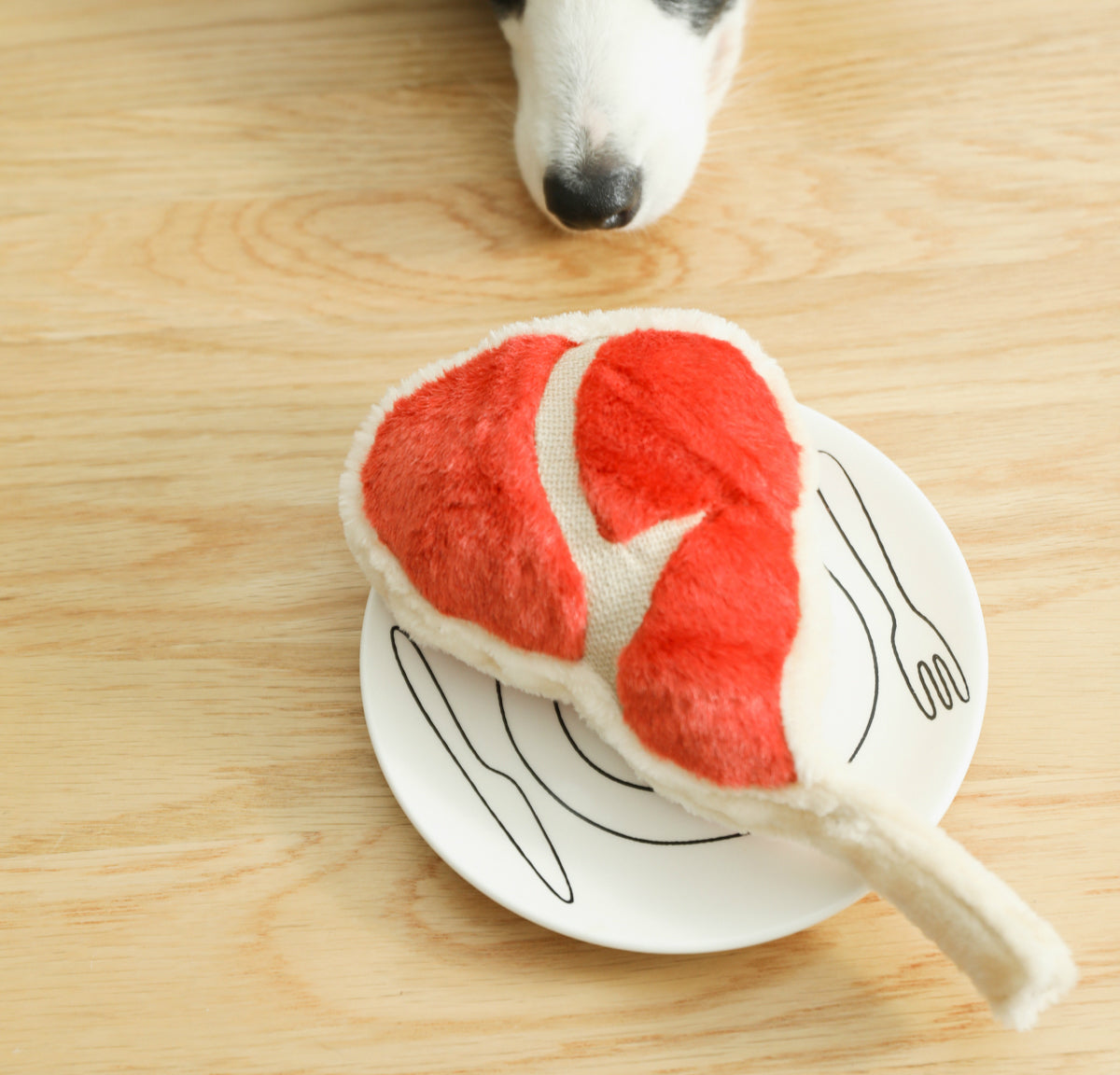 One red-and-white fuzzy steak-shaped plush toy on a white plate with black utensil drawings, dog nose above.