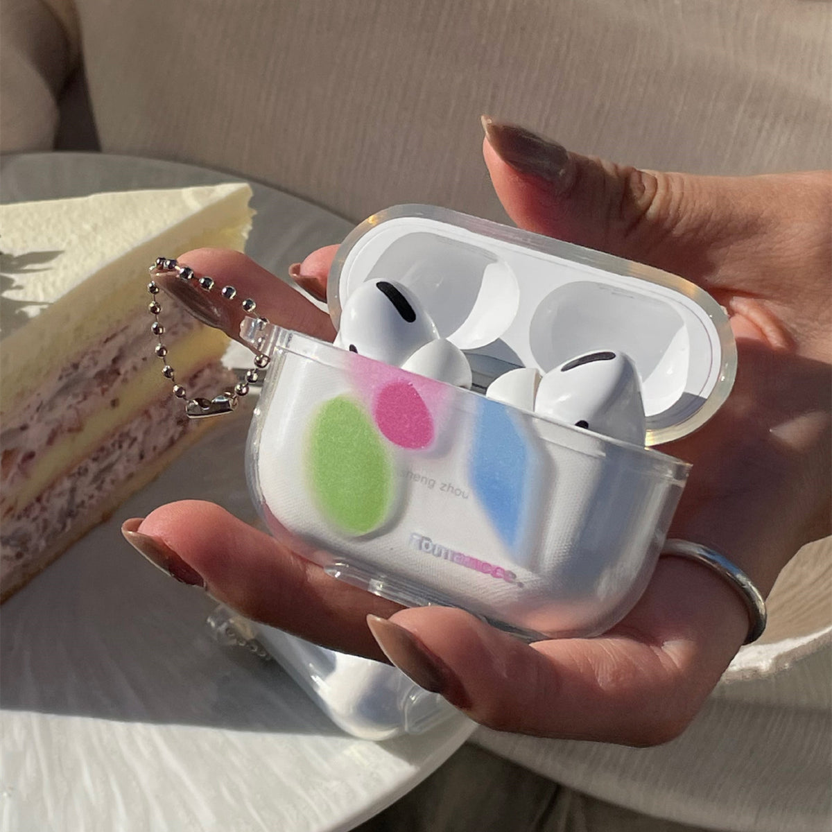 Clear protective case held in hand, open with two white earbuds, green pink blue spots and silver ball chain.