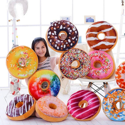 11 large colorful donut plush pillows with assorted frostings and sprinkles around a seated woman.