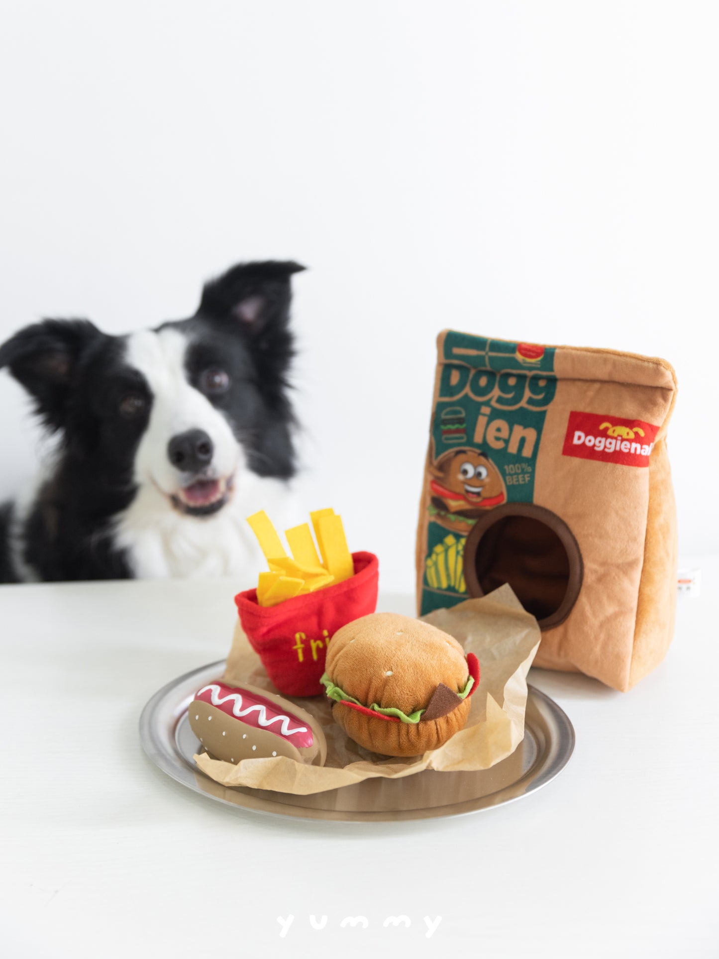 Border collie behind a tray of plush food: burger, hot dog, red fries and a tan Doggienal plush bag.