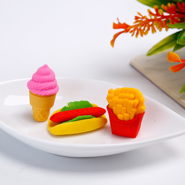 Three food erasers on white plate: pink ice-cream cone, yellow hot dog with green lettuce, red box of fries.
