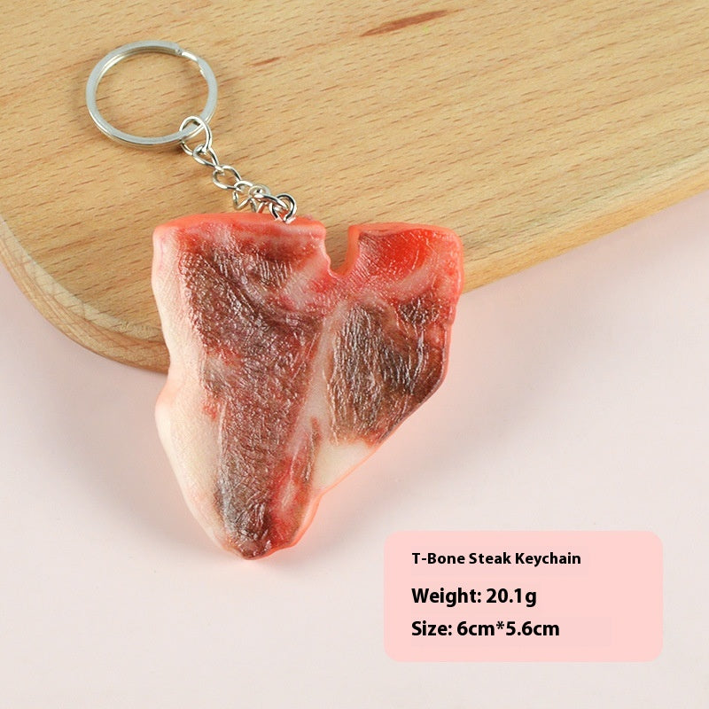 One realistic T-bone steak keychain (red, brown, white fat texture) with silver chain and ring on a board.