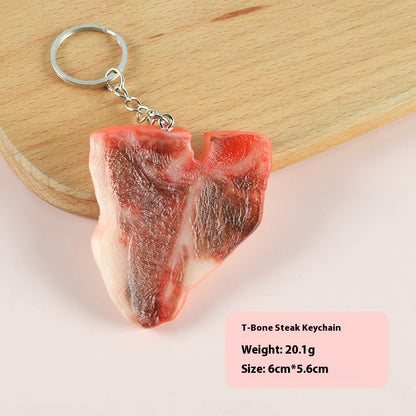 One realistic T-bone steak keychain (red, brown, white fat texture) with silver chain and ring on a board.