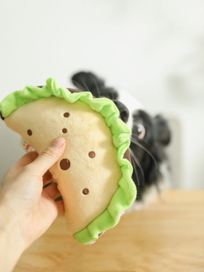 Hand holding one beige plush taco toy with green ruffled edge and brown dot details; blurred black dog behind.