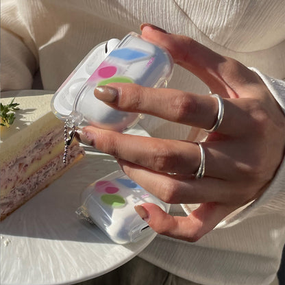 Hand with silver rings holds clear earbud case with pink blue green dots; another case and cake slice.