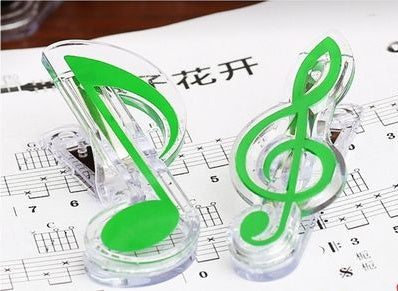 Two green musical-note bag clips (eighth note and treble clef) with clear plastic bases on sheet music.