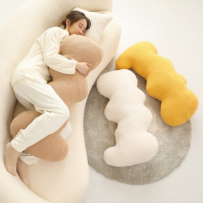 Three cloud-shaped fuzzy cushions in beige, cream and mustard, one hugged by a woman on a sofa.