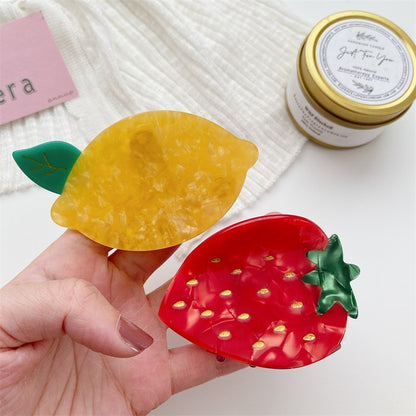 Two fruit hair clips held in hand: yellow lemon with green leaf; red strawberry with gold seeds.