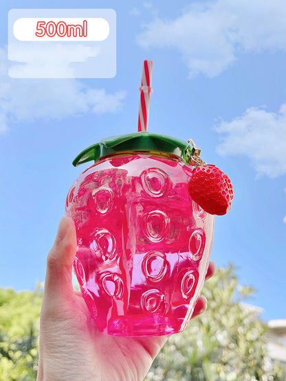 Hand holding translucent hot-pink strawberry-shaped 500ml cup with green lid, striped straw and red charm.