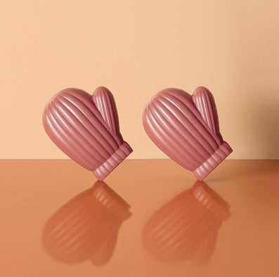 Two dusty-rose mitten-shaped bag clips with vertical ribbing and cuff detail, reflected on a peach surface.