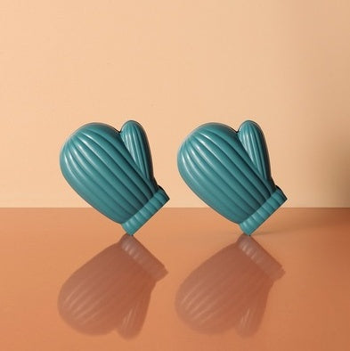 Two teal ribbed mitten-shaped bag clips tilted on a reflective surface against a beige background.