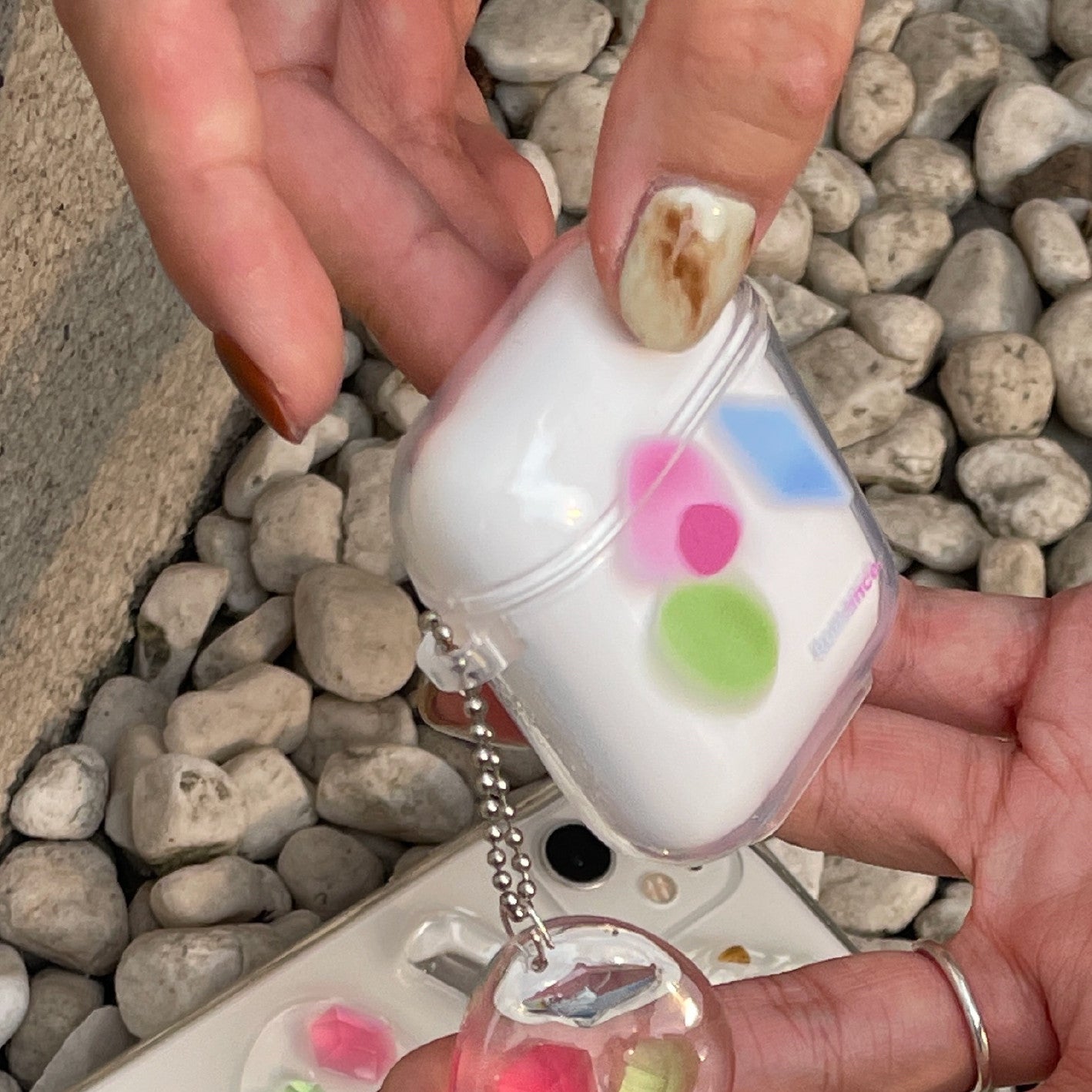 One white AirPods case in clear shell with pink, green and blue dots and silver ball-chain charm.