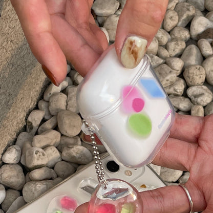 One white earbud case in a clear shell with pink and green dots and a clear charm, held over gray pebbles.