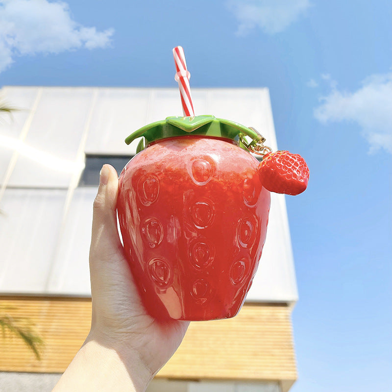 One glossy red strawberry cup with green leaf lid, striped straw and small strawberry charm, held in hand.