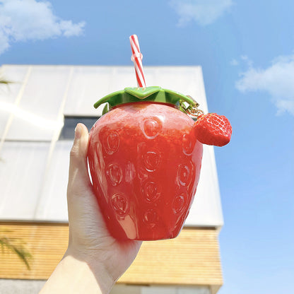 One glossy red strawberry cup with green leaf lid, striped straw and small strawberry charm, held in hand.