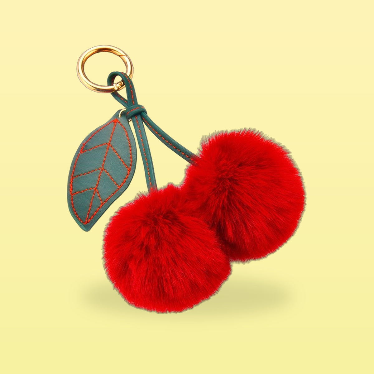 Fluffy red pom-pom cherry bag charm with a dark green stitched leaf and gold clip on a butter yellow background