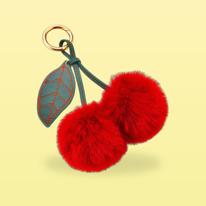 Fluffy red pom-pom cherry bag charm with a dark green stitched leaf and gold clip on a butter yellow background