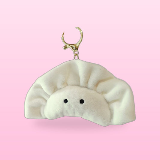White plush dumpling keychain with two dot eyes and a gold clasp, on a pink background