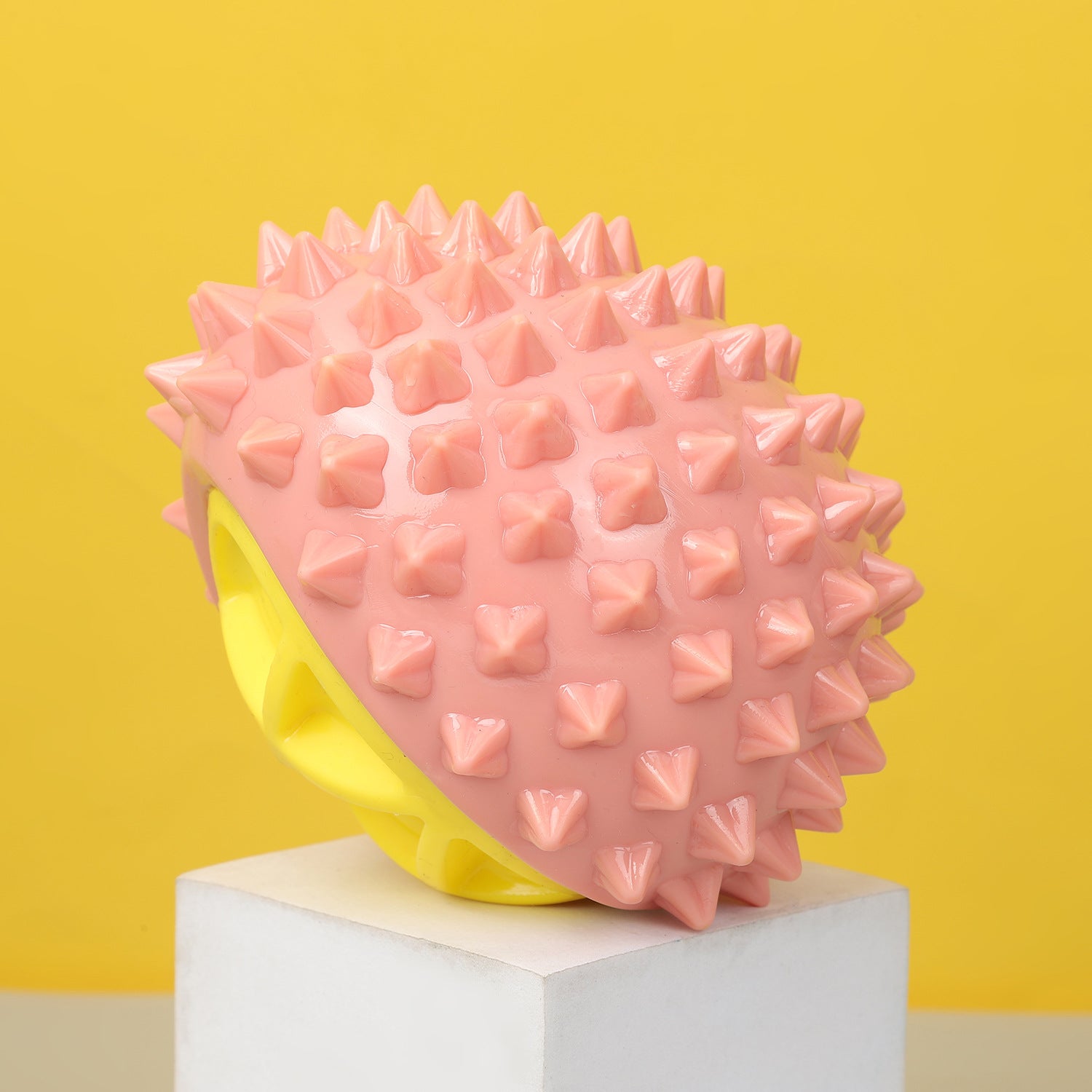 One pink spiky rubber chew toy with a yellow hollow base on a white pedestal against a yellow background.