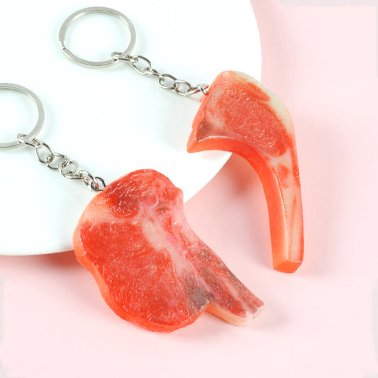 Two red-and-white faux pork chop keychains with silver chains on a white plate, pink background.