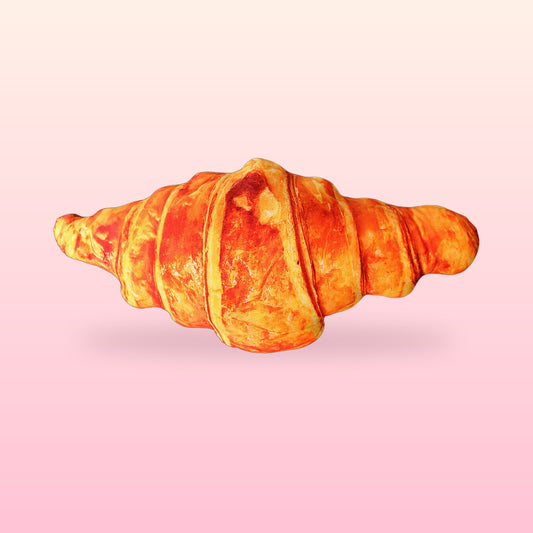 Giant croissant hug pillow with a golden flaky bakery print on a soft pink background