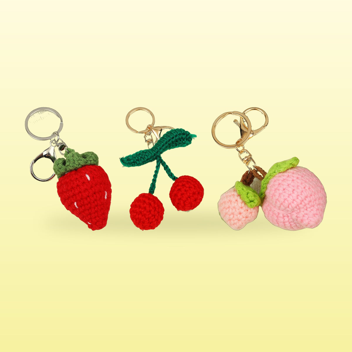 Three crochet fruit keychains, a strawberry, cherries and two peaches, on a butter yellow background